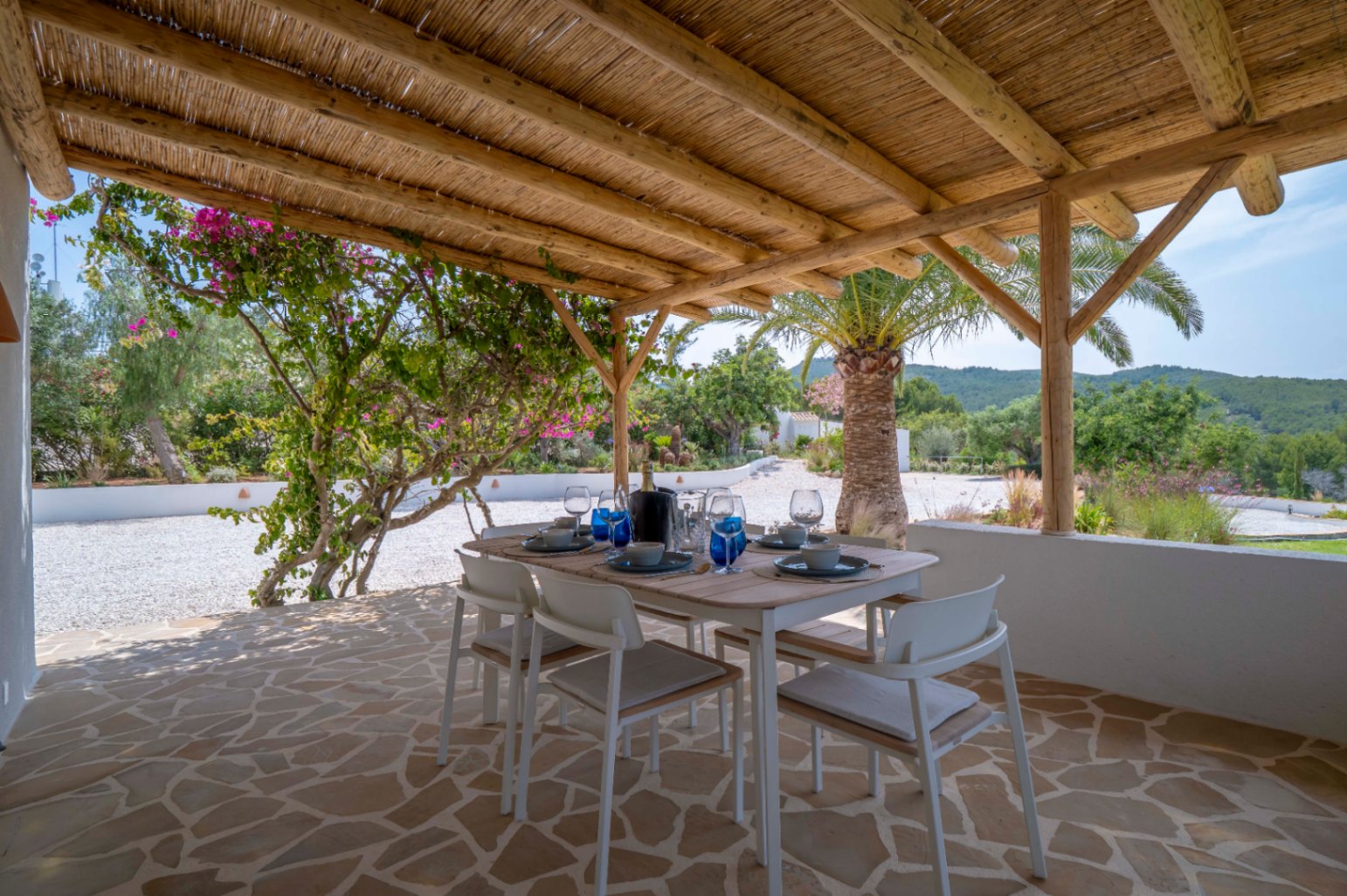 Salle à manger extérieure couverte avec toit de chaume, sol en pierre irrégulière et vue sur le jardin méditerranéen.