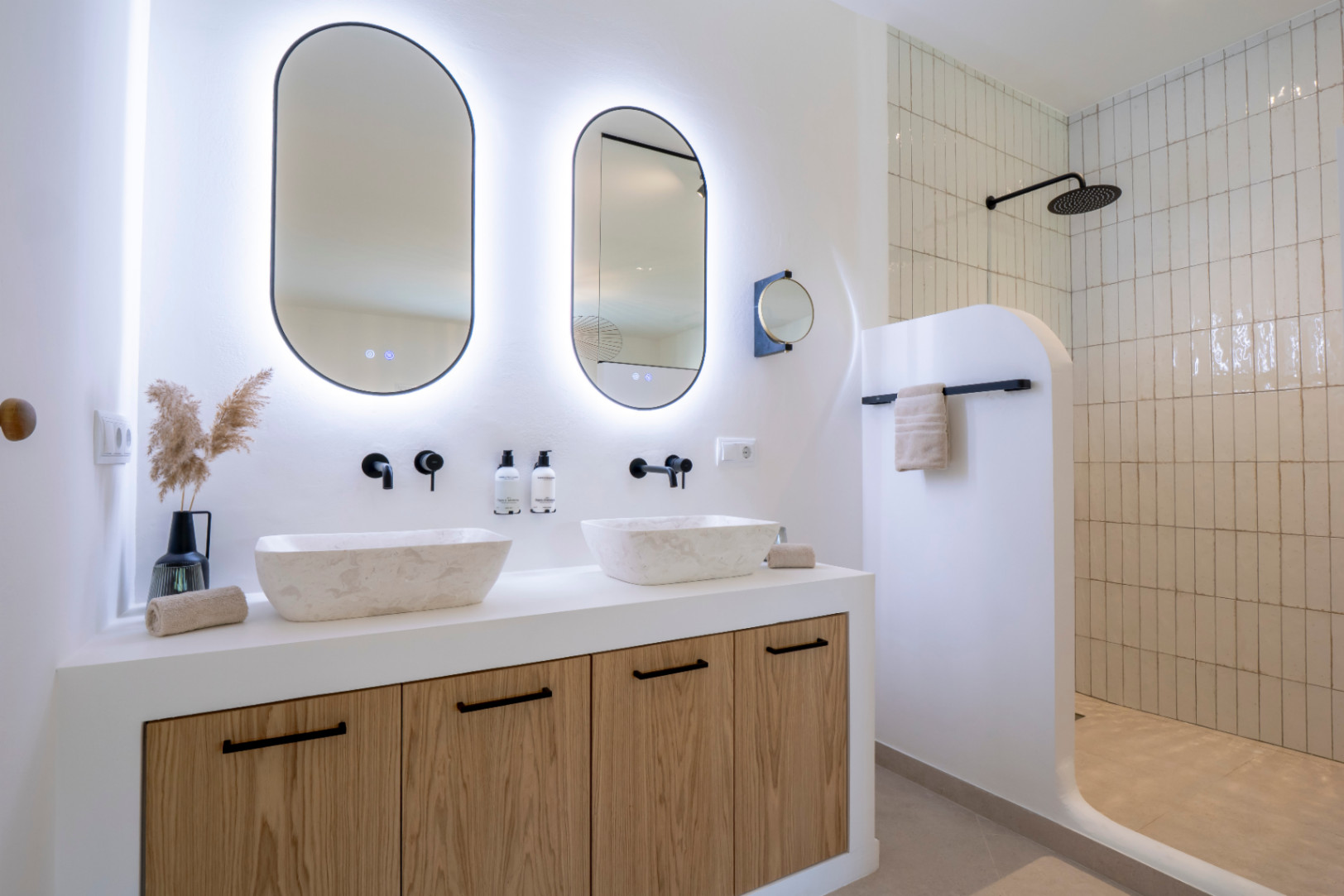 Salle de bain élégante avec lavabos en pierre, miroirs ovales lumineux et douche carrelée verticalement.