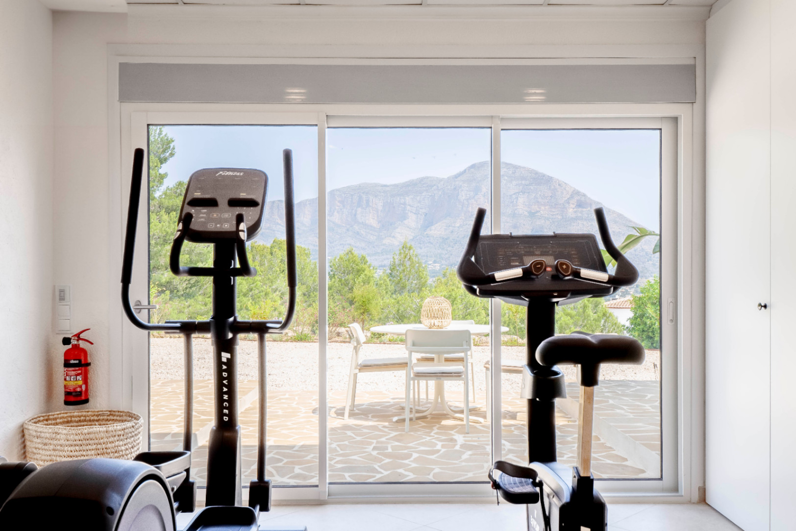 Salle de sport lumineuse avec vue sur la montagne, équipée d'un vélo et d'un elliptique.