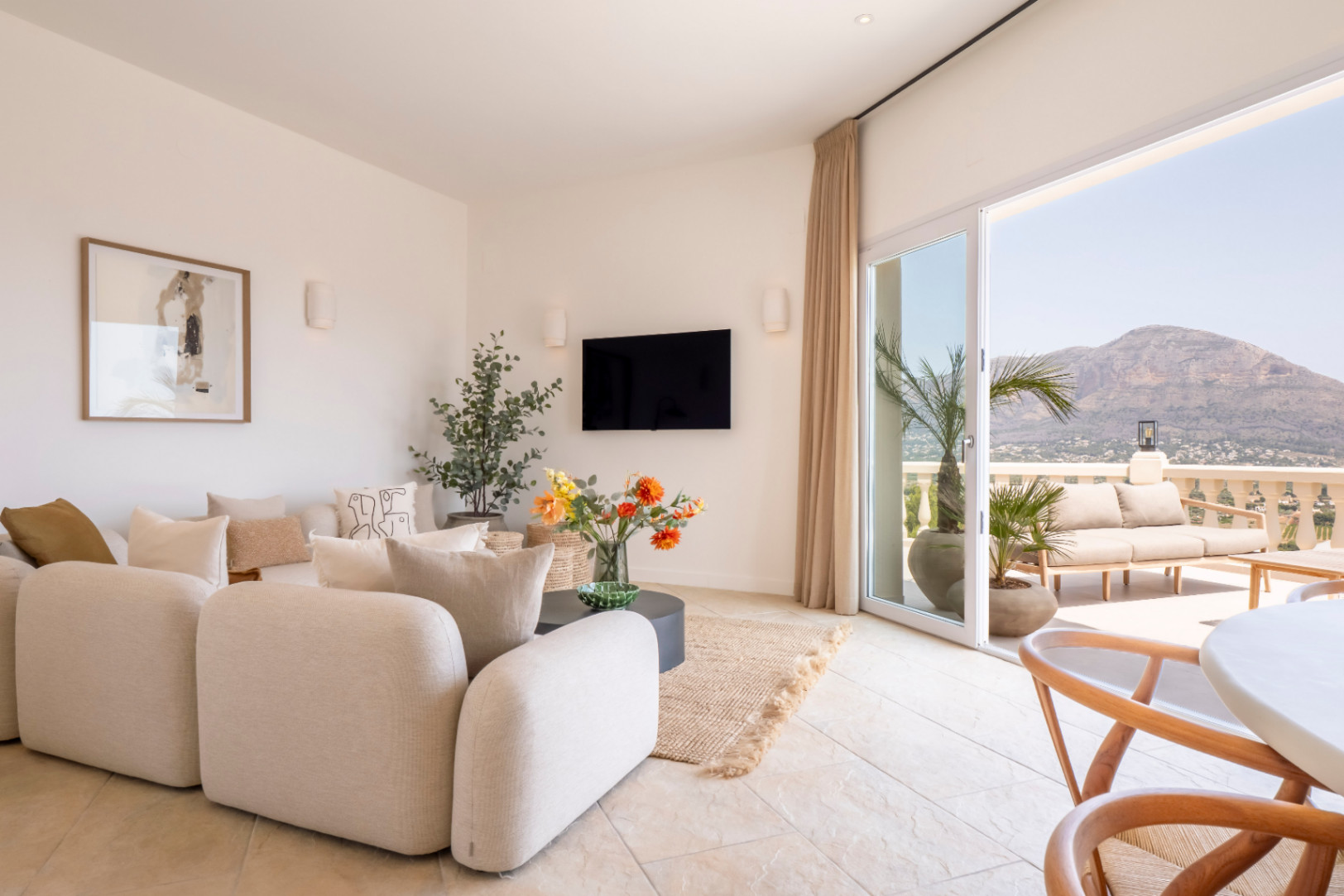 Salon lumineux avec accès terrasse. Canapé modulable beige, TV et décor floral. Vue sur la montagne à Jávea.