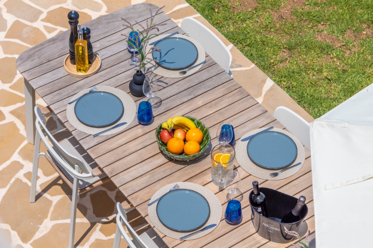 Table à manger extérieure avec assiettes bleues, verrerie et coupe de fruits, prête pour un repas au jardin.
