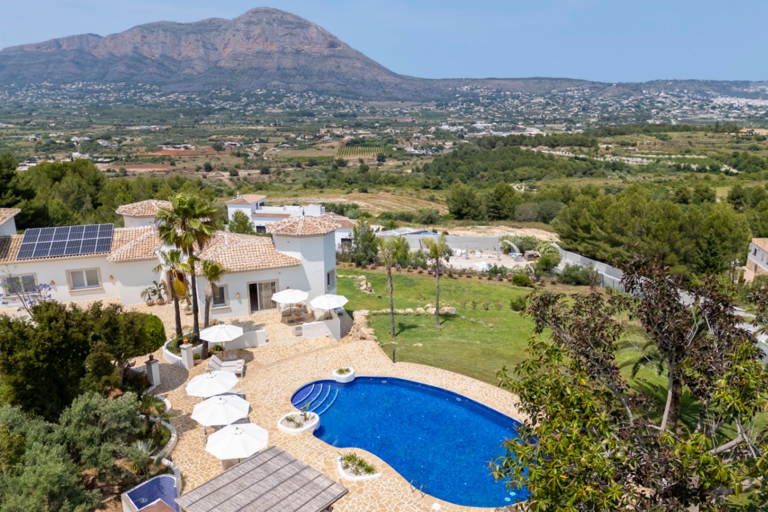 Vue aérienne de la villa avec piscine bleue, terrasses avec parasols blancs et jardin méditerranéen avec vue sur la montagne.