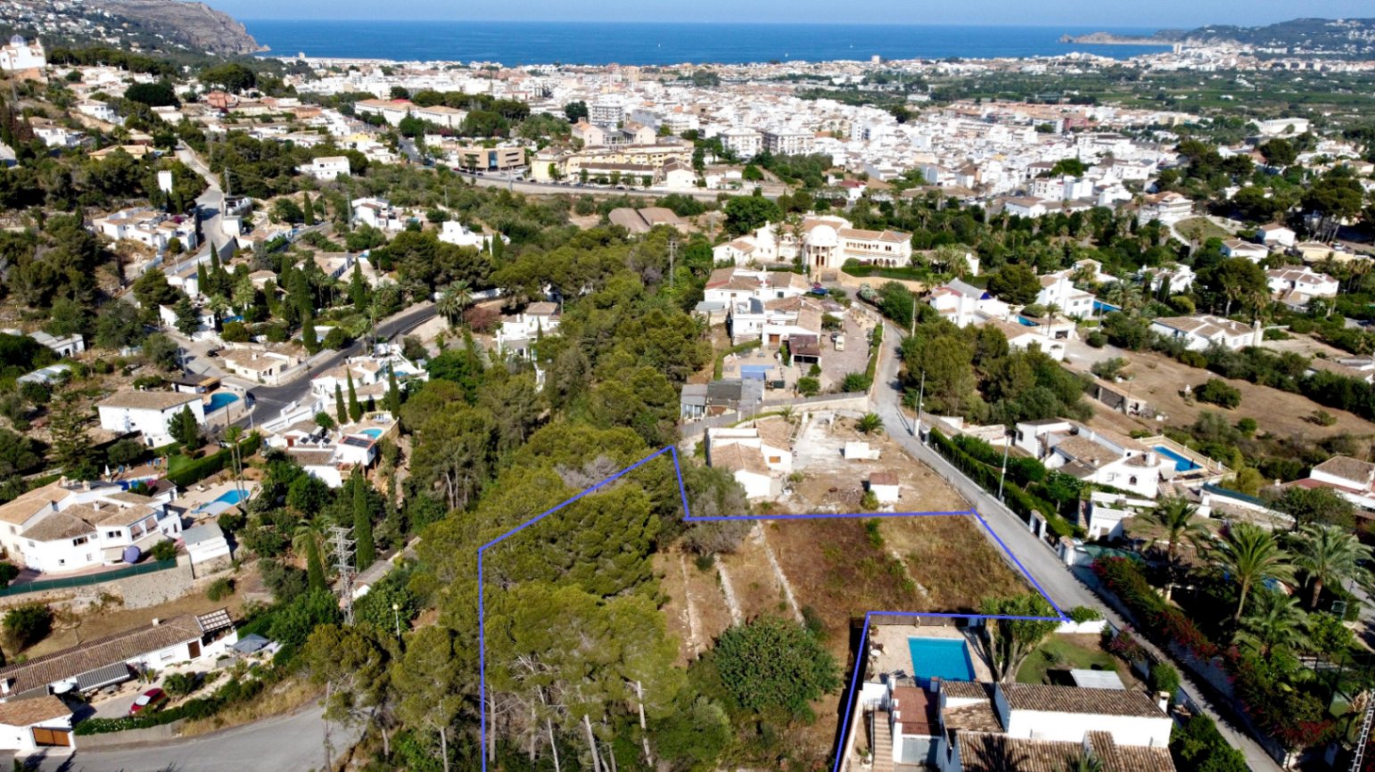 Vue aérienne de la parcelle de 1 908 m² délimitée en bleu, comprenant une pinède et offrant une vue panoramique sur la mer et Jávea.
