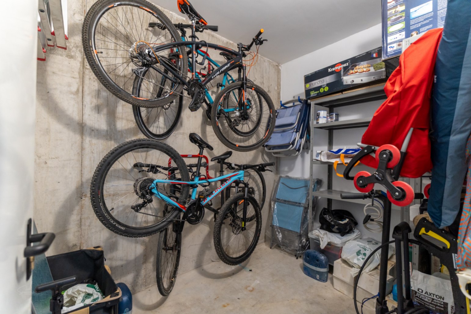 Cellier privé avec murs en béton brut, équipé de supports muraux pour vélos et d'étagères métalliques pour un rangement optimisé du matériel.