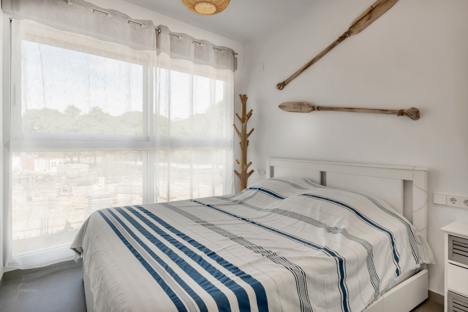 Chambre lumineuse avec lit double, décoration nautique comprenant des rames en bois et un porte-manteau artisanal près de la grande fenêtre.