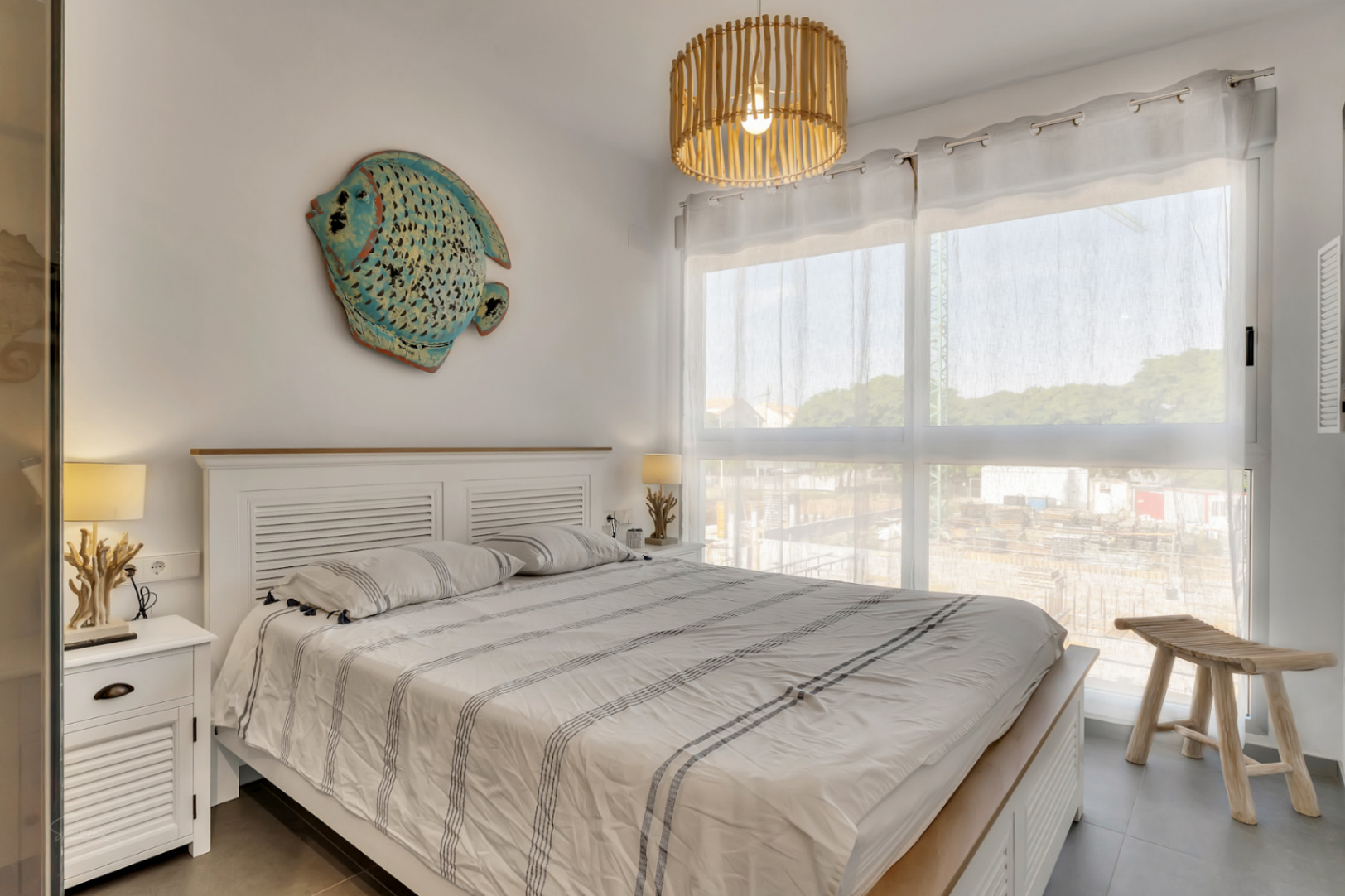 Chambre lumineuse avec lit double, tête de lit en bois blanc et décoration marine. Une grande fenêtre illumine l'espace, complétée par un luminaire en bambou.