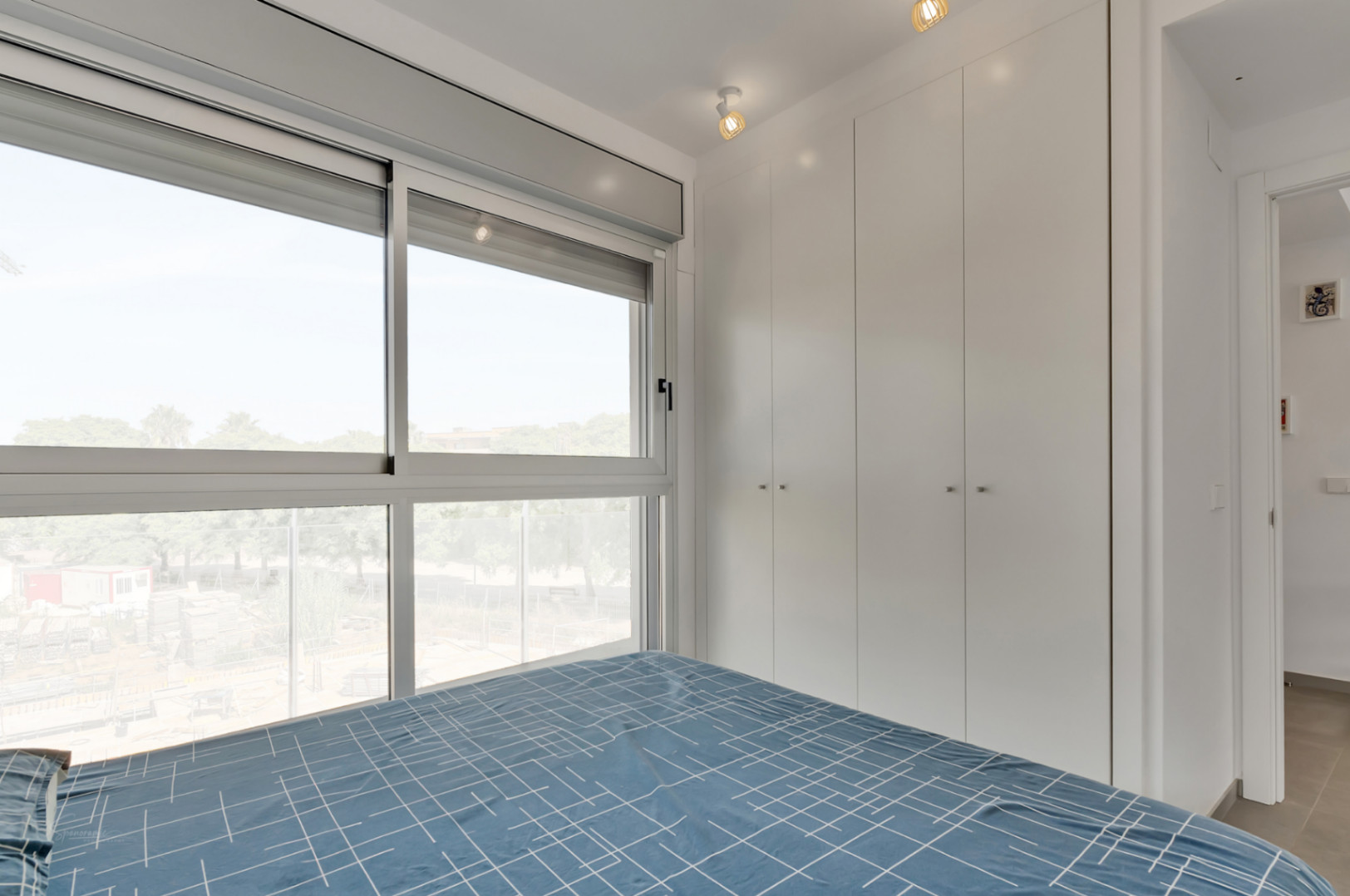 Chambre lumineuse avec placards intégrés blancs, grandes fenêtres coulissantes offrant une vue dégagée et éclairage moderne par spots.