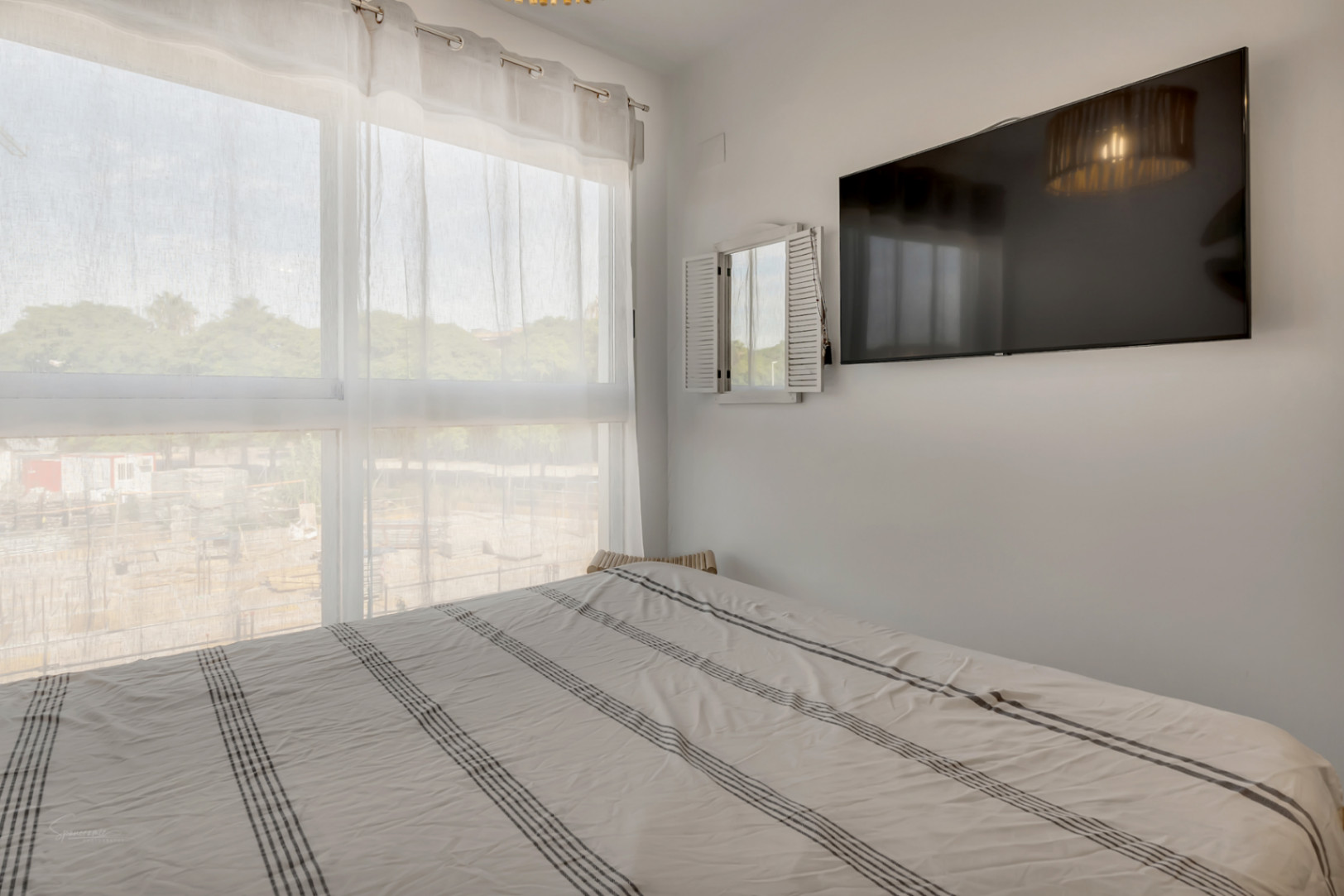 Chambre lumineuse avec une grande fenêtre, rideaux translucides, lit double à rayures et télévision à écran plat fixée au mur blanc.