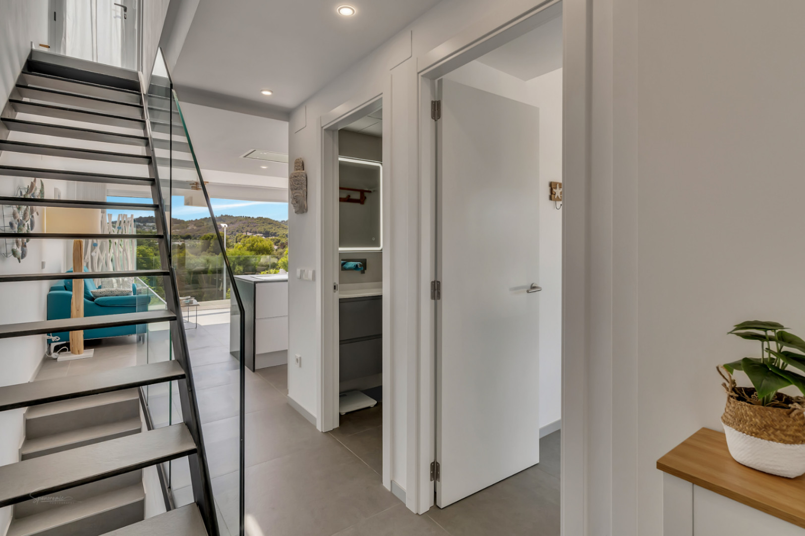 Couloir moderne avec escalier à marches suspendues et garde-corps en verre. Vue sur une salle de bain contemporaine et le salon lumineux ouvert sur l'extérieur.