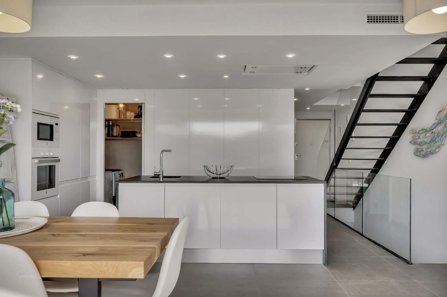 Cuisine moderne minimaliste avec îlot central, placards blancs laqués et buanderie. Un escalier design en métal noir et verre sépare la cuisine de l'espace repas en bois.