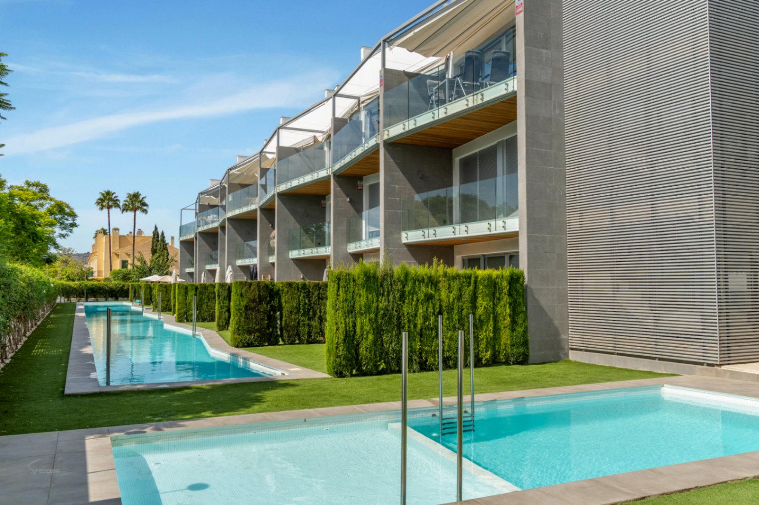 Façade moderne avec balcons vitrés et finitions en bois, surplombant un espace commun avec deux piscines et des haies de cyprès taillées.