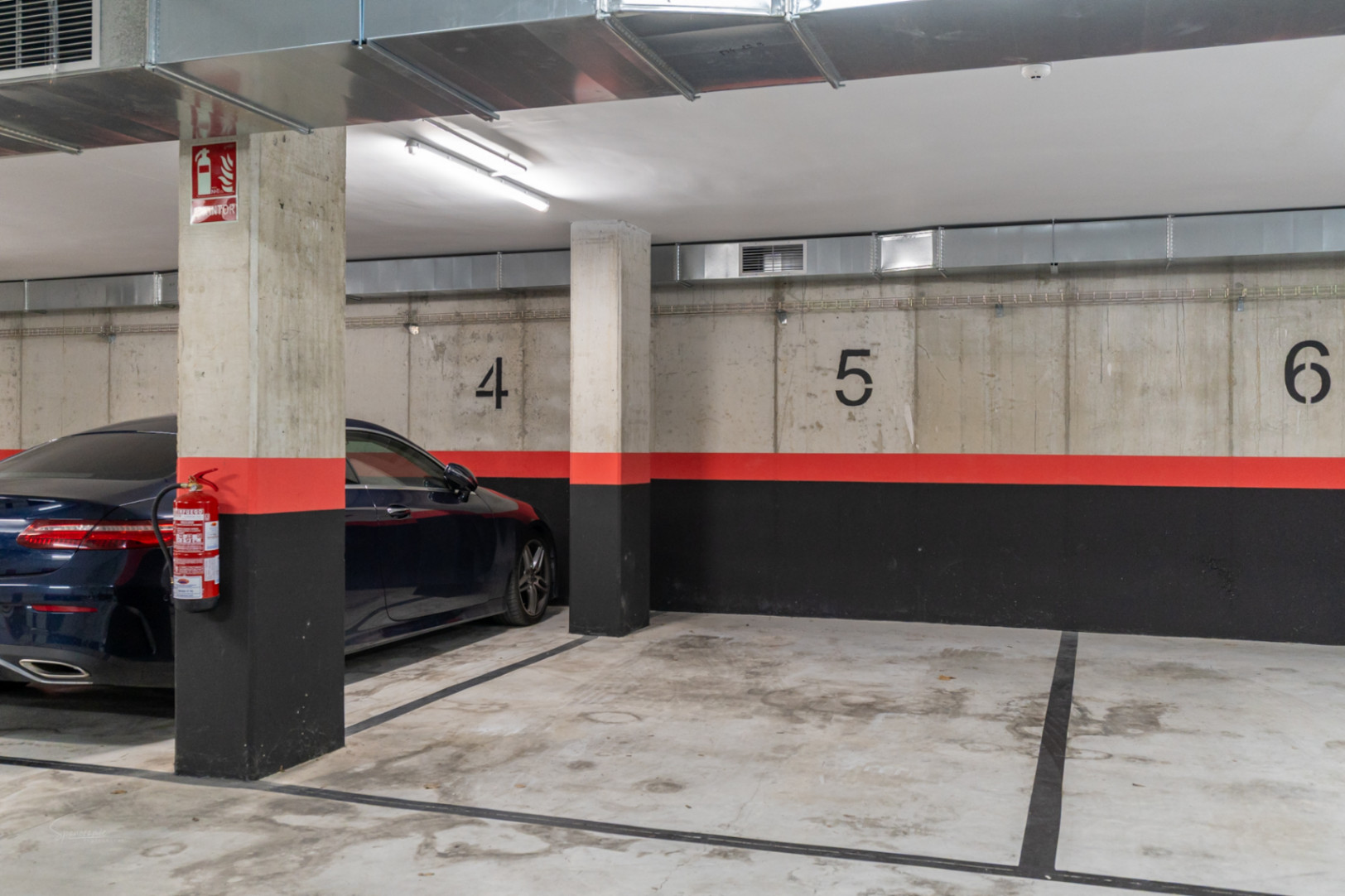 Place de parking souterraine numérotée avec murs en béton brut, conduits de ventilation apparents et dispositifs de sécurité incendie.