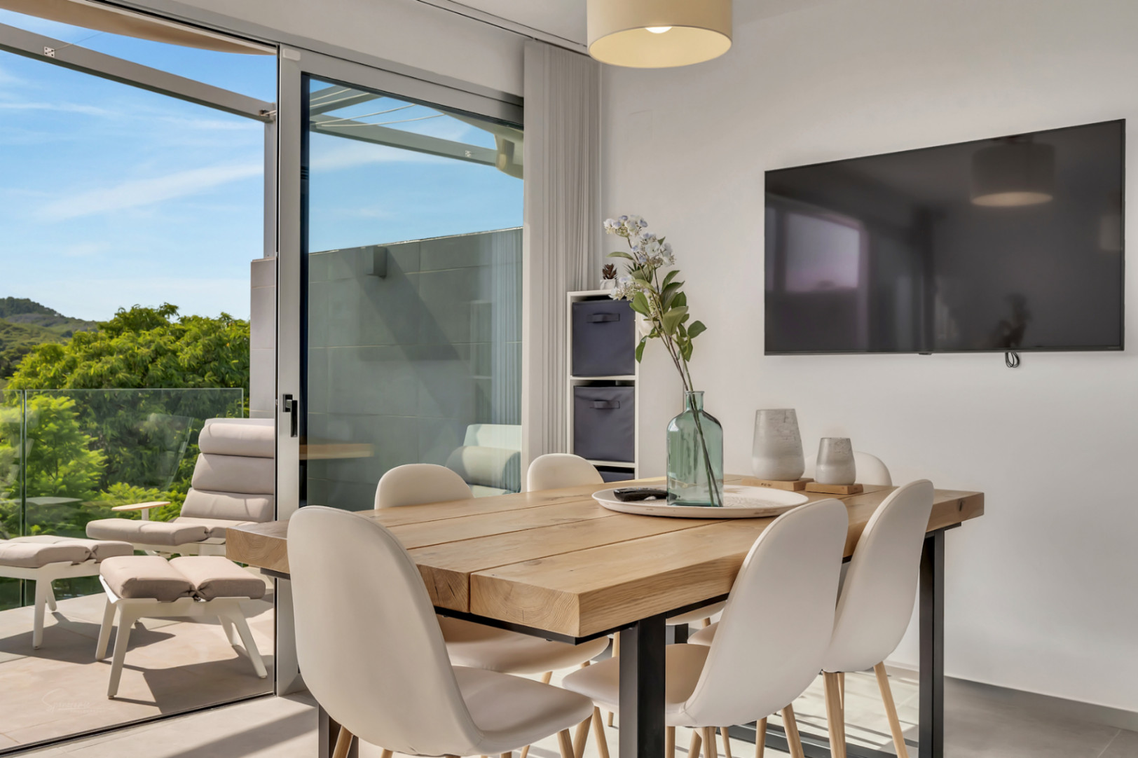 Salle à manger lumineuse avec table en bois massif, s'ouvrant sur un balcon équipé de fauteuils et offrant une vue sur la nature environnante.
