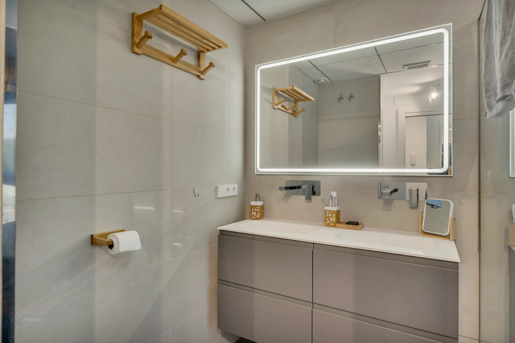 Salle de bain moderne avec meuble double vasque suspendu gris et miroir rétroéclairé par LED. Robinetterie murale chromée et accessoires élégants en bois naturel.