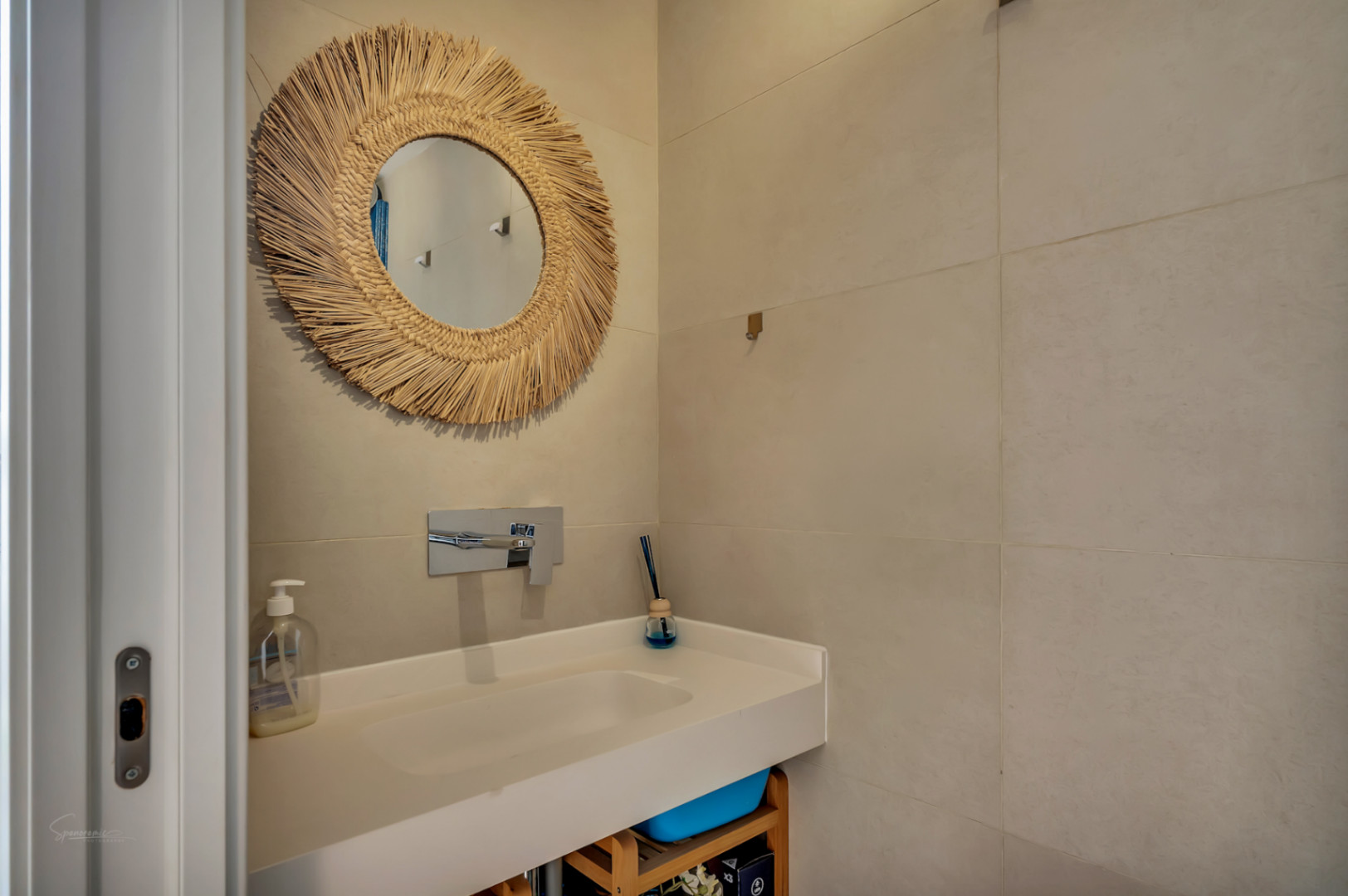 Salle de bains moderne avec vasque intégrée blanche, robinetterie murale chromée et miroir décoratif en fibres naturelles sur un carrelage grand format ton sable.