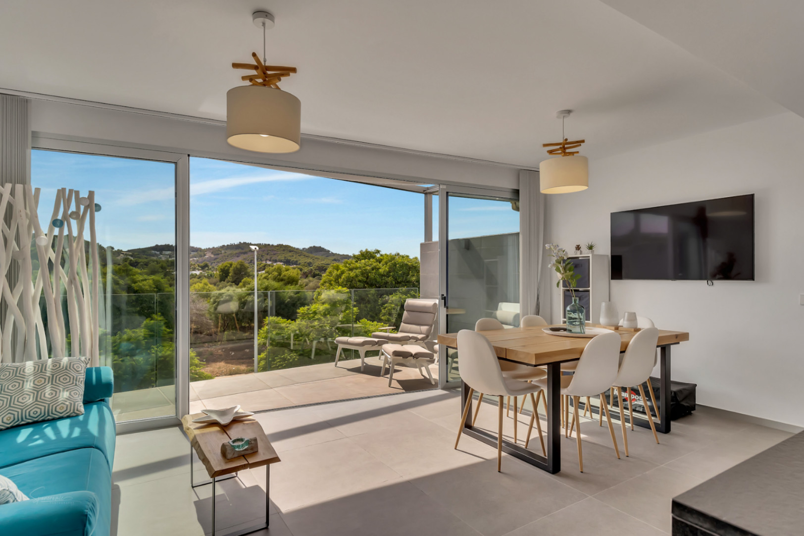 Salon-salle à manger lumineux avec de grandes baies vitrées coulissantes ouvrant sur un balcon avec vue sur la nature. Table en bois, chaises modernes et carrelage grand format.