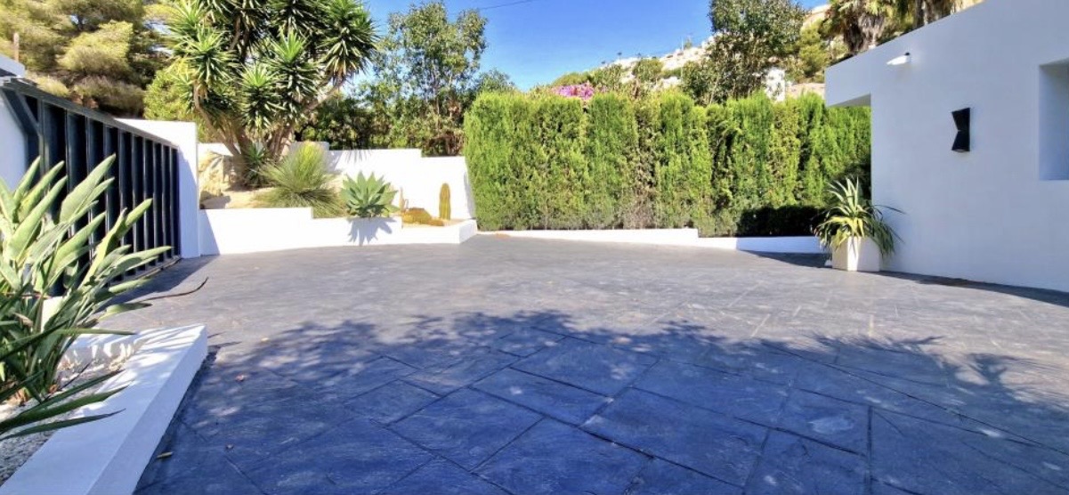 Aire de stationnement avec revêtement en béton imprimé sombre, entourée de murs blancs, d'une haie de cyprès et d'un jardin méditerranéen avec des yuccas.