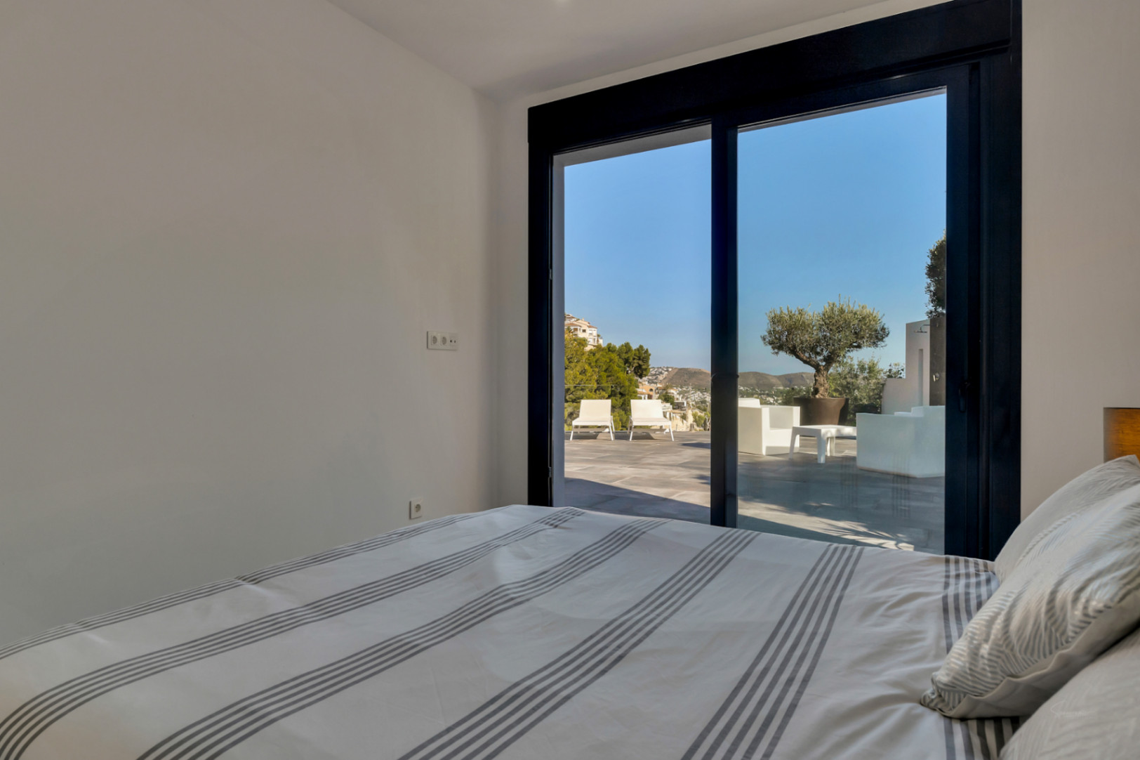 Chambre avec accès direct à la terrasse par une baie vitrée noire. L'espace extérieur comprend du mobilier design, un olivier et une vue dégagée sur les montagnes.