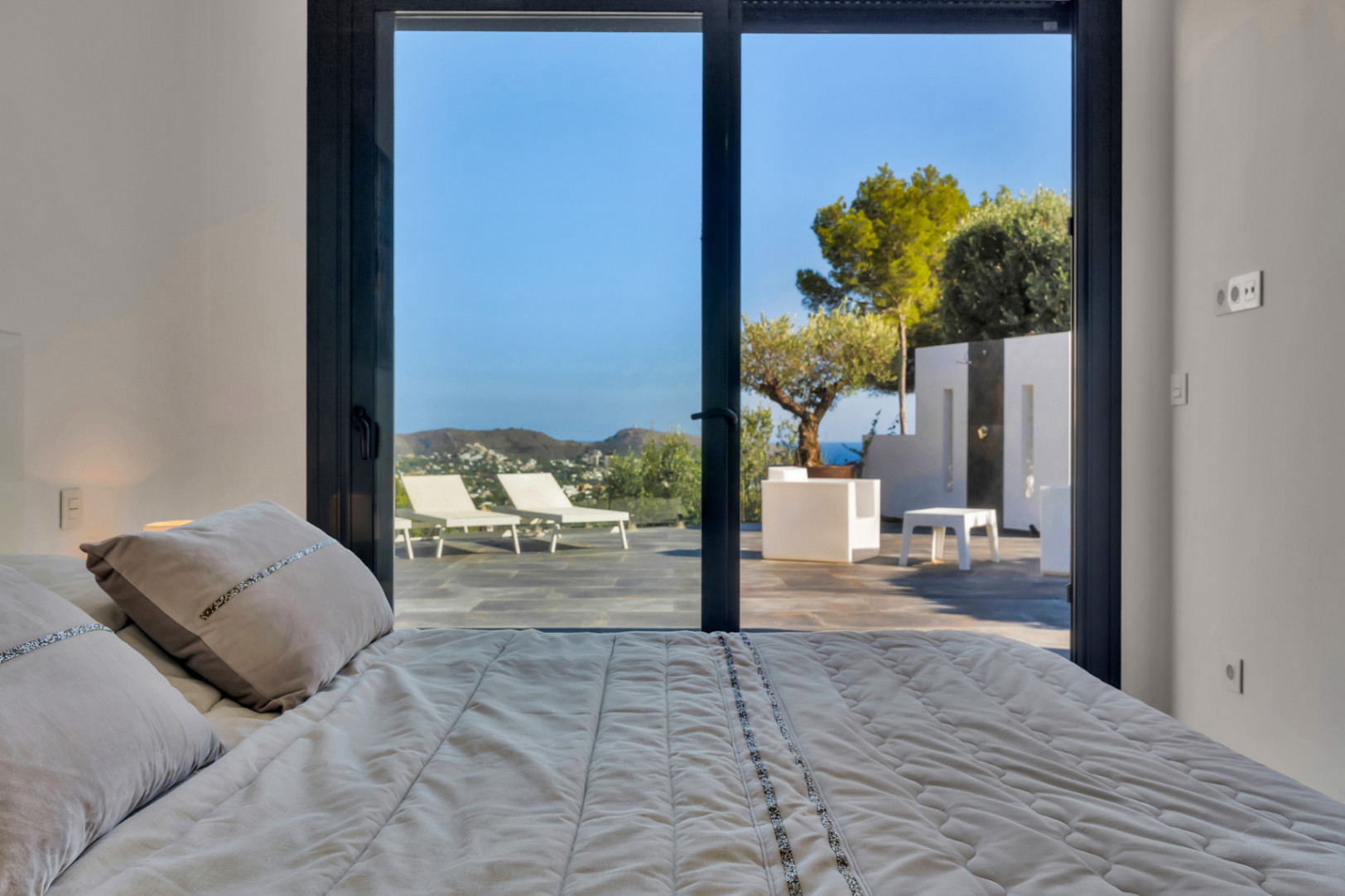 Chambre avec baies vitrées coulissantes ouvrant sur une terrasse en pierre, équipée de mobilier blanc, d'une douche extérieure et vue sur la mer.