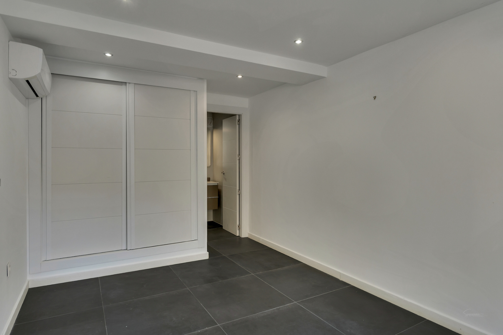 Chambre fonctionnelle avec placard intégré blanc, sol en grès cérame gris foncé, climatisation et accès direct à la salle de bain attenante.