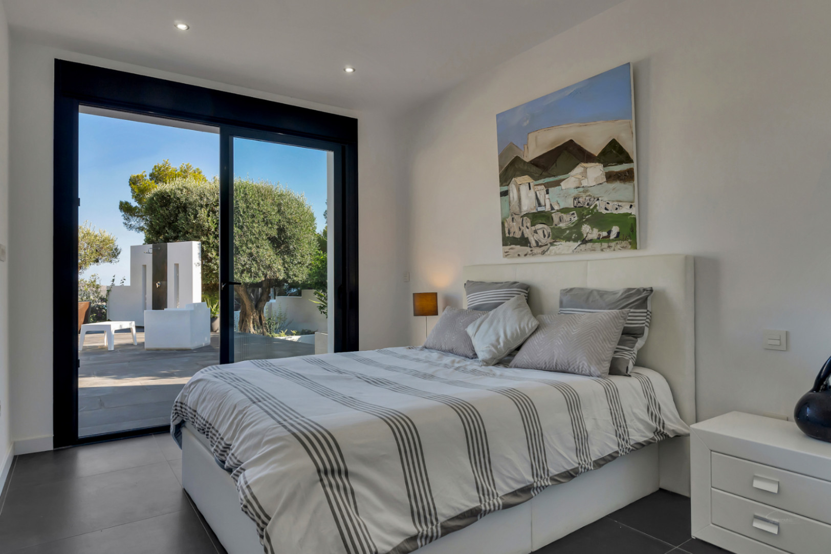 Chambre moderne avec accès direct à la terrasse, baies vitrées à cadre noir, sol en céramique foncée et éclairage encastré au plafond.