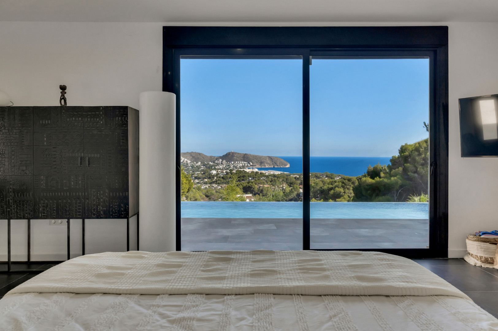 Chambre principale avec vue panoramique sur la mer et la piscine à débordement. Comprend des baies vitrées, un mobilier de style et un sol sombre.