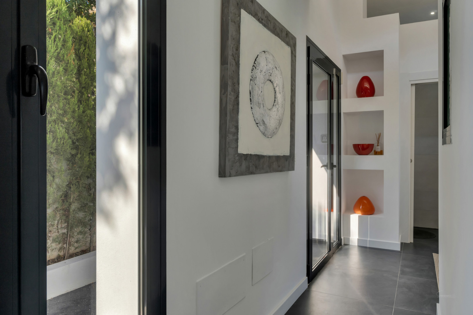 Couloir moderne avec sol en grès cérame foncé, niches décoratives intégrées et menuiseries noires laissant entrer la lumière naturelle et la vue sur le jardin.