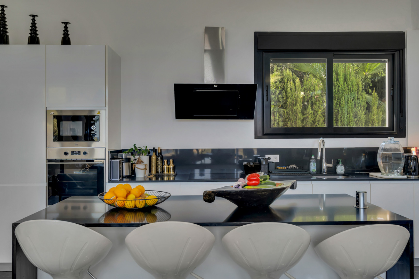 Cuisine à aire ouverte avec îlot noir, tabourets design blancs et fenêtre offrant une vue sur le jardin méditerranéen.