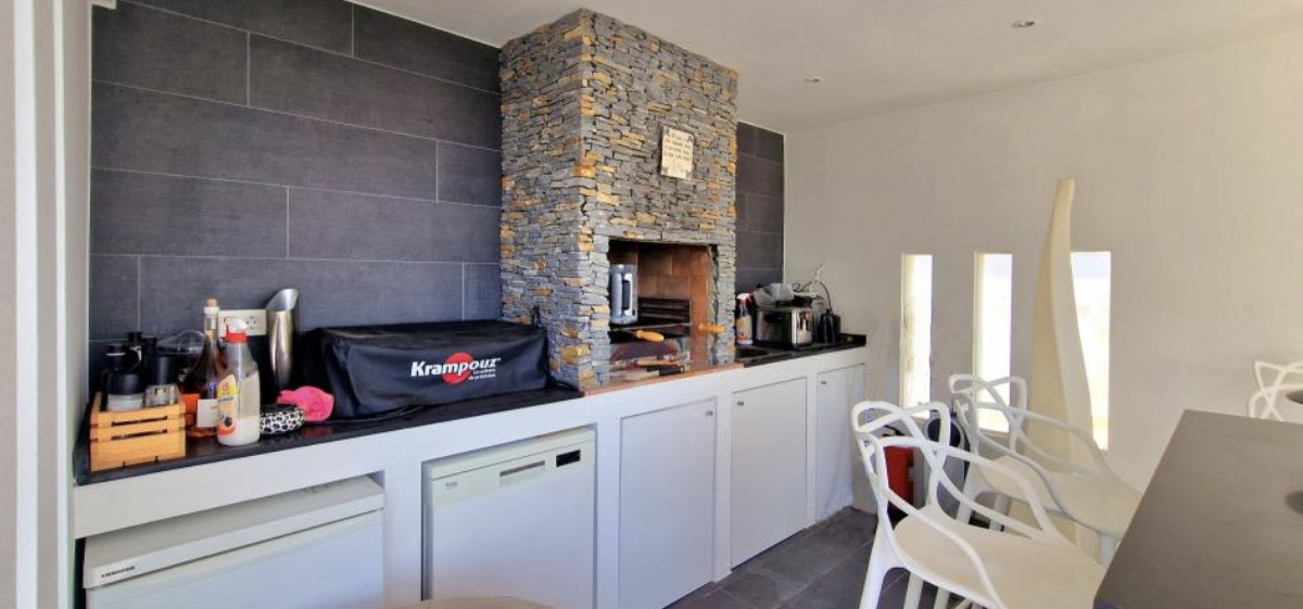 Cuisine d'été équipée d'un barbecue en pierre naturelle, d'un plan de travail noir et d'appareils intégrés sous des meubles blancs épurés.