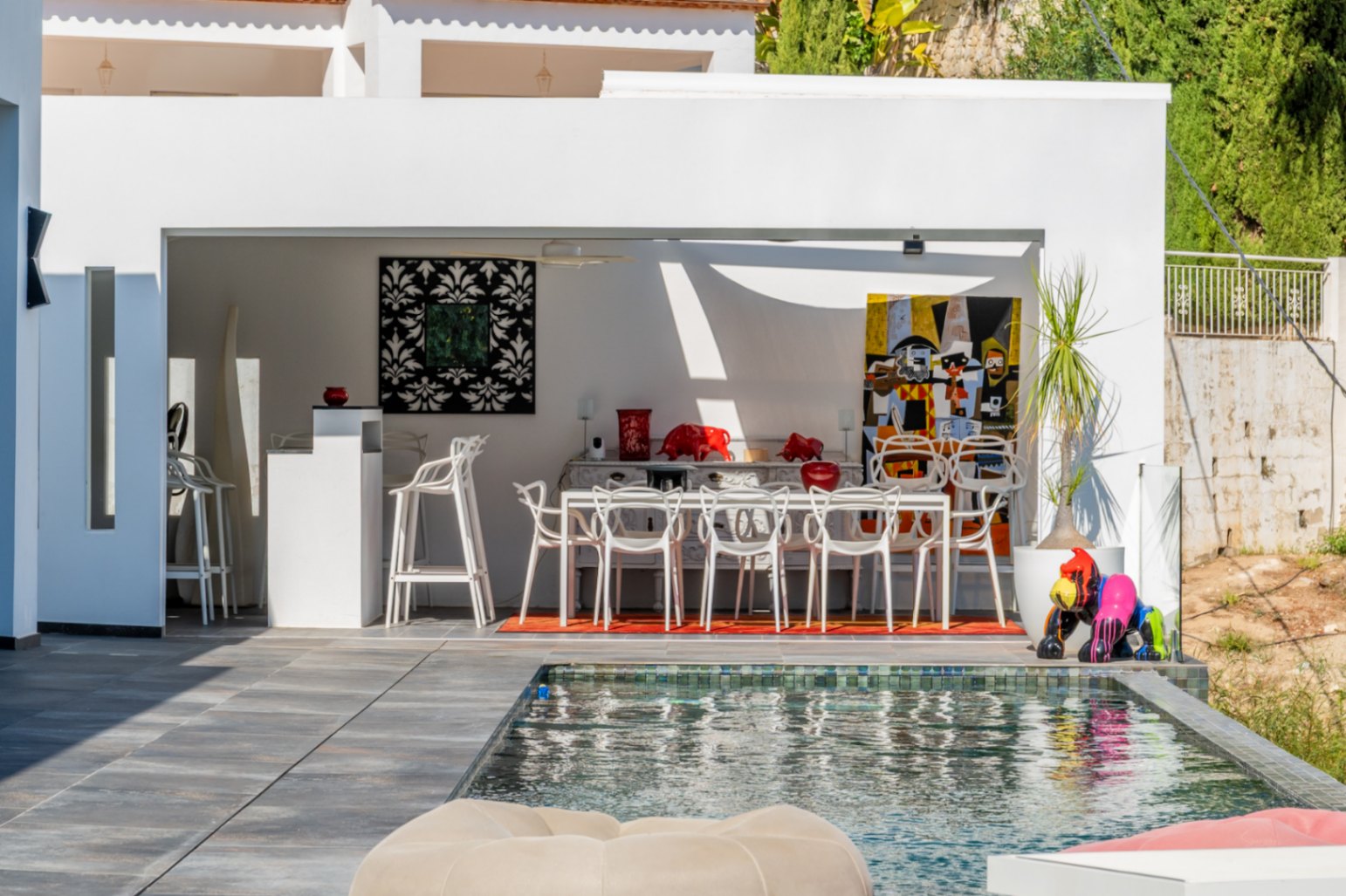 Cuisine d'été et salle à manger extérieure avec mobilier design blanc et œuvres d'art colorées, située au bord d'une piscine aux reflets sombres.