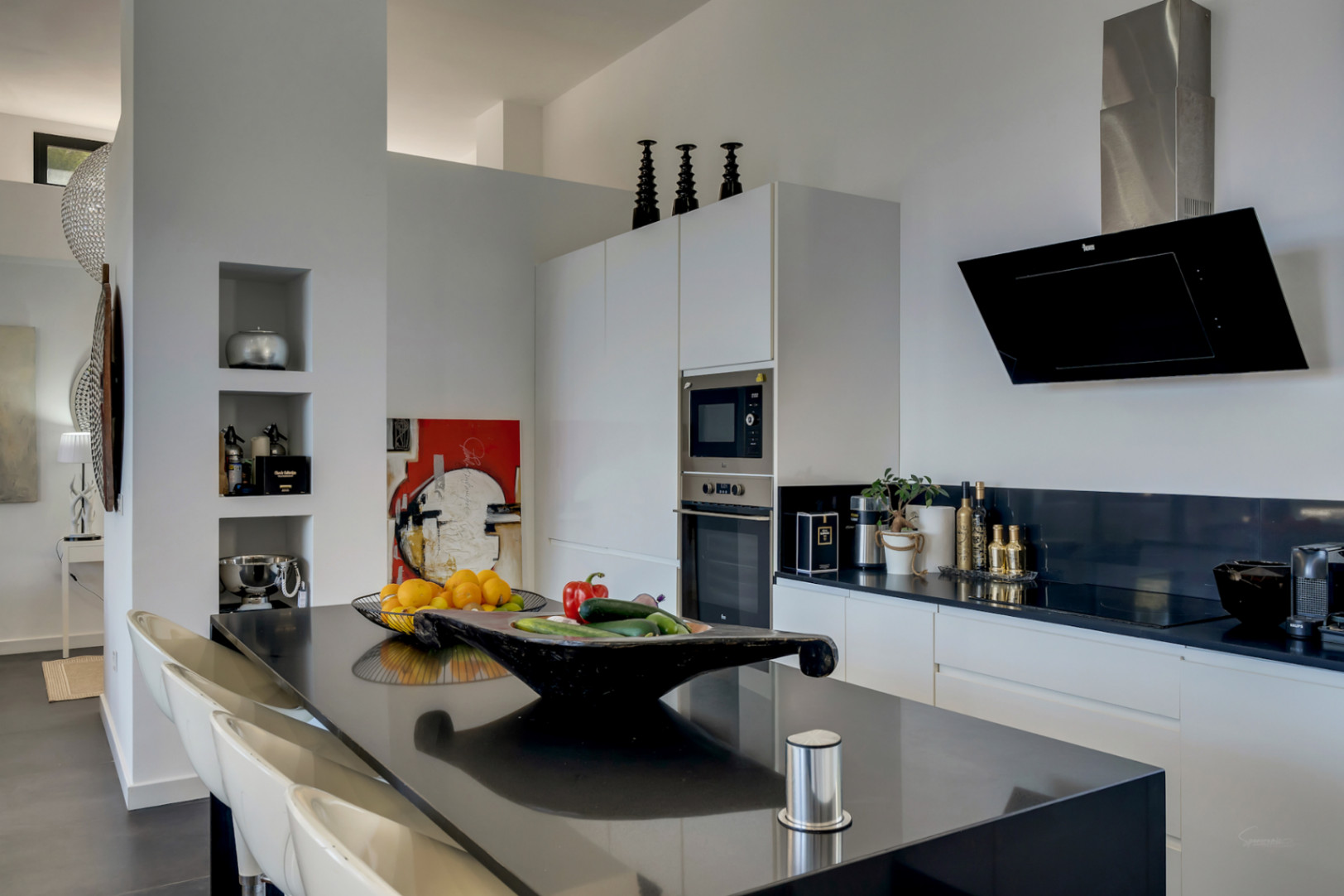 Cuisine minimaliste avec îlot central noir, électroménagers intégrés et placards blancs épurés. Elle dispose d'une hotte en verre noir et de niches de rangement murales intégrées.