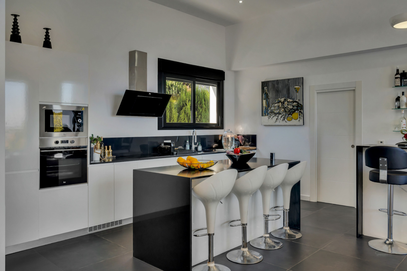 Cuisine ouverte avec îlot central noir, tabourets design blancs et électroménager encastré. Le contraste entre le blanc et le noir souligne l'esthétique moderne.