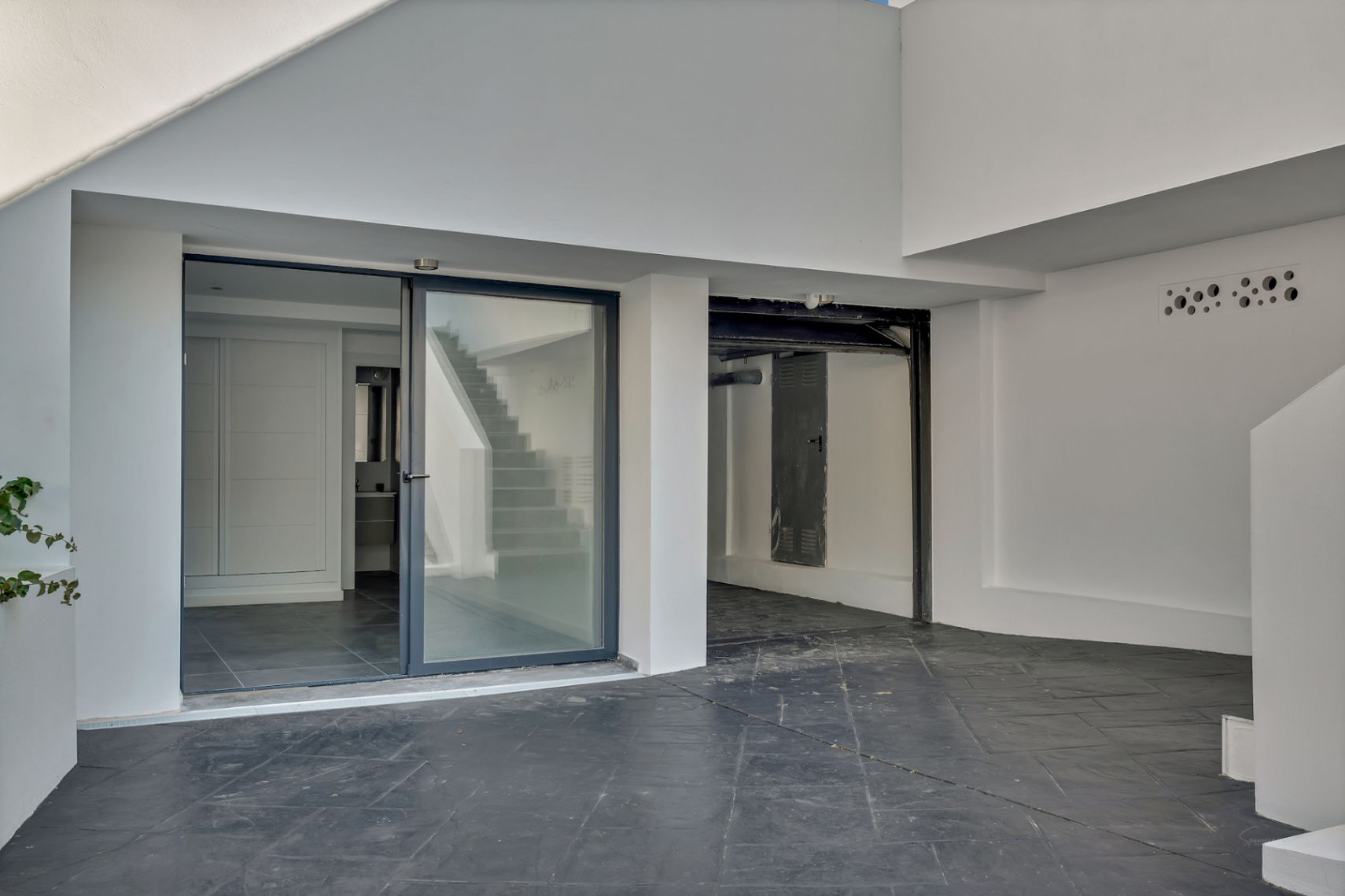 Entrée du niveau inférieur avec porte vitrée coulissante, sol en dalles sombres et espace de garage ou rangement intégré à l'architecture blanche épurée.