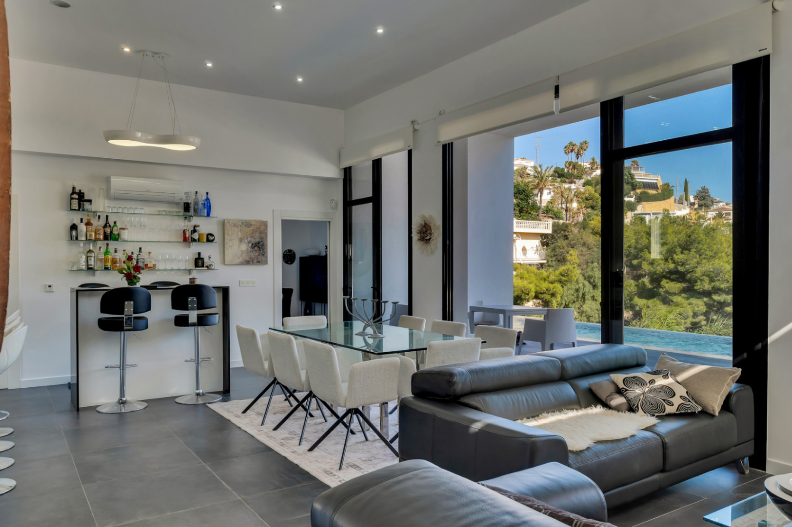 Espace de vie ouvert comprenant un salon, une salle à manger et un bar, avec de grandes baies vitrées offrant une vue sur la piscine et les collines environnantes.