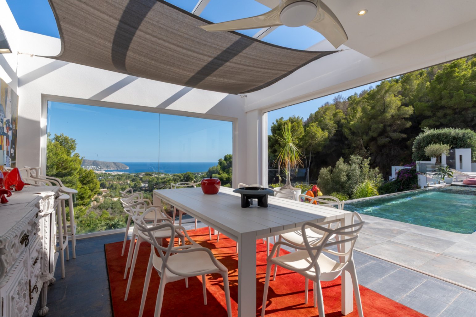 Espace dînatoire extérieur vitré avec vue panoramique sur la mer, mobilier design, ventilateur de plafond et accès direct à la piscine à débordement.