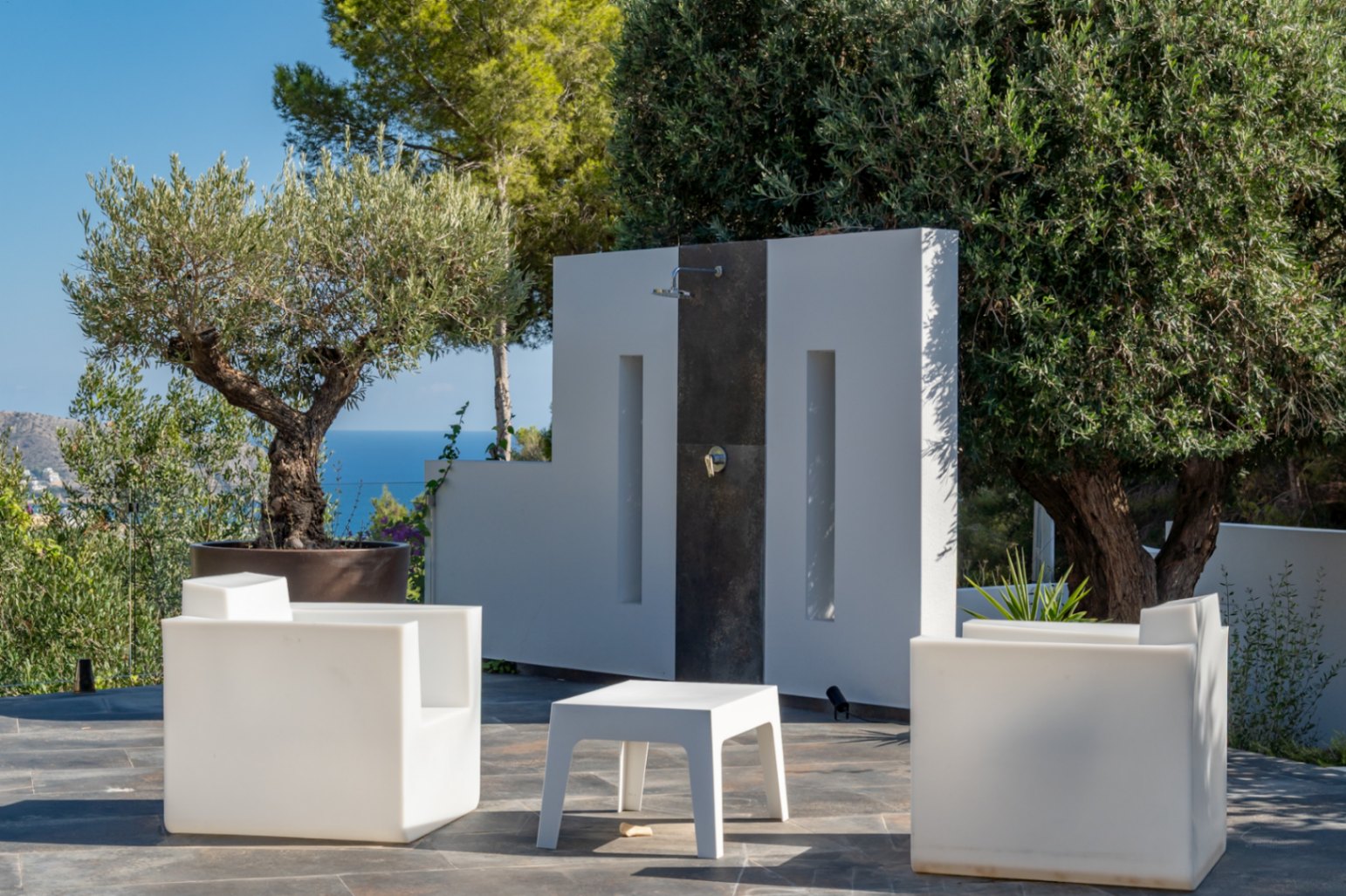 Espace douche extérieure au design épuré avec mobilier blanc, sol en pierre naturelle et vue dégagée sur la mer et la végétation locale.