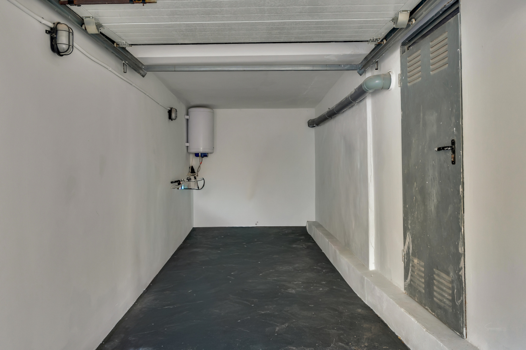 Garage ou espace de rangement fonctionnel avec sol sombre, murs blancs, chauffe-eau électrique et porte latérale métallique ventilée.