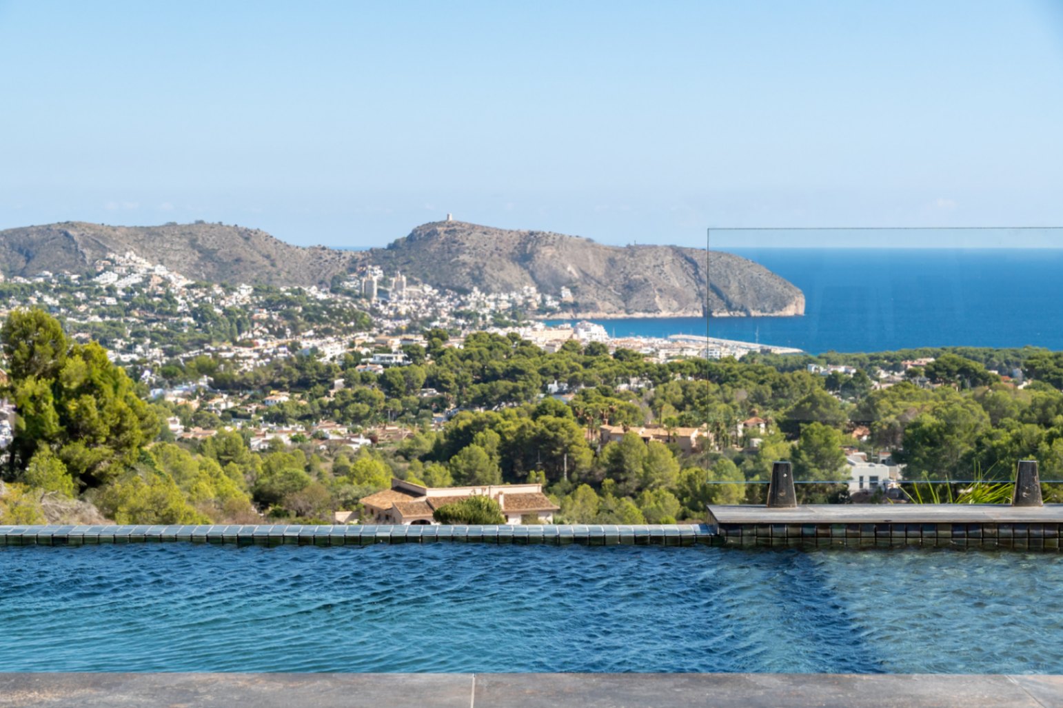 Piscine à débordement avec mosaïque bleue et garde-corps en verre, offrant une vue panoramique sur la mer Méditerranée, le Cap d'Or et la pinède environnante.