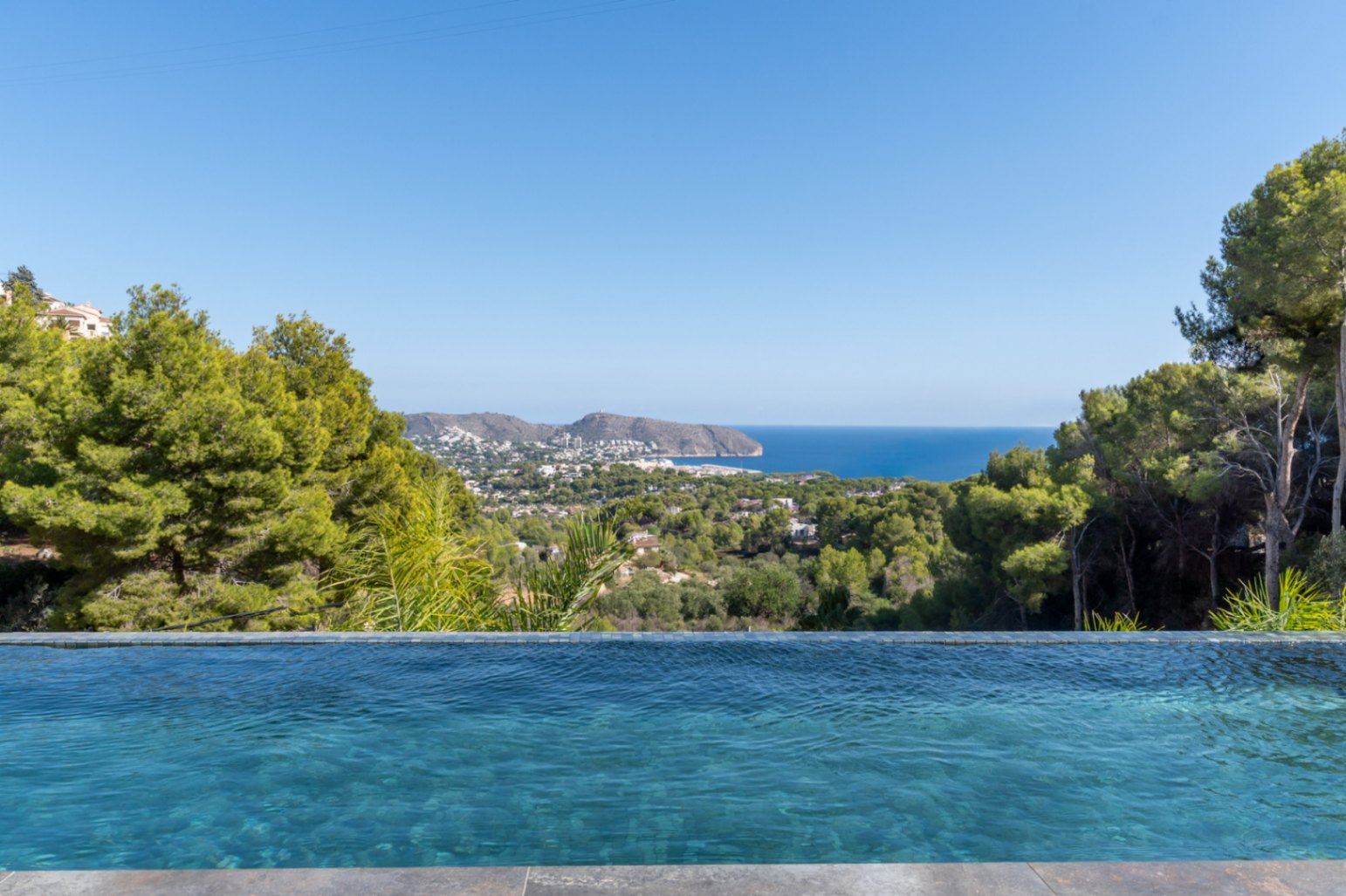 Piscine à débordement avec mosaïque bleue offrant une vue panoramique sur la mer Méditerranée, la côte de Moraira et la pinède environnante.