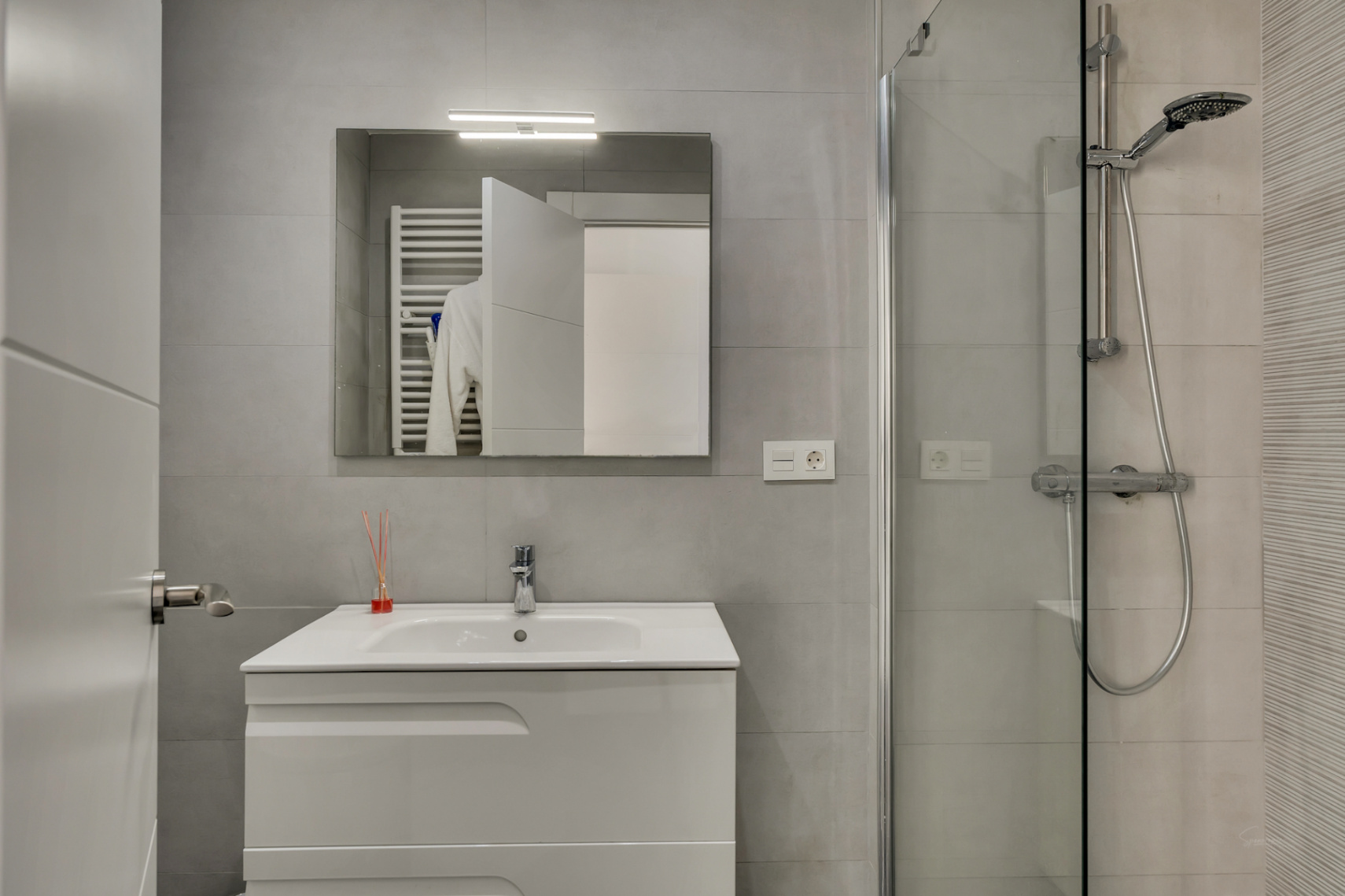 Salle de bain moderne avec carrelage gris, meuble vasque blanc, miroir éclairé et douche à l'italienne avec paroi en verre et robinetterie chromée.