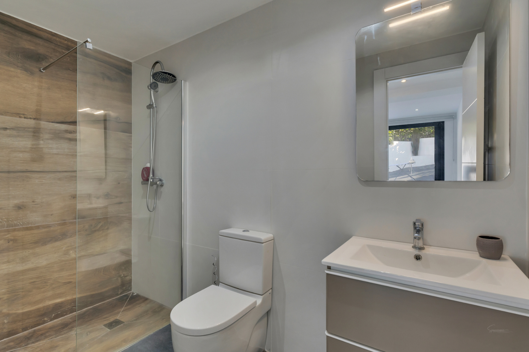 Salle de bain moderne avec douche à l'italienne en grès effet bois, paroi vitrée, toilettes blanches et meuble vasque avec miroir à éclairage intégré.