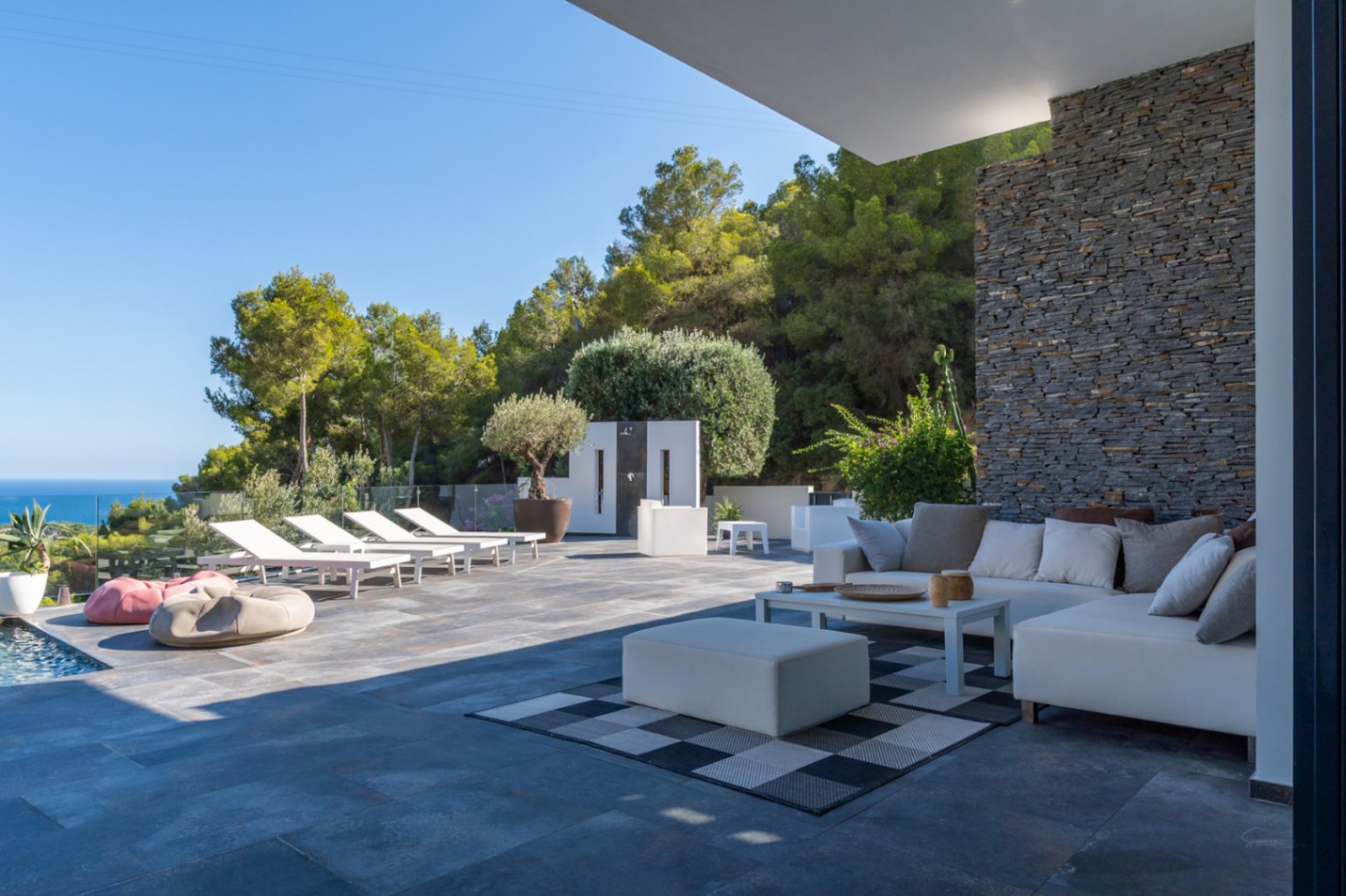 Salon d'été avec canapés modulables blancs, mur en pierre naturelle et terrasse spacieuse offrant un solarium et une vue panoramique sur la mer.