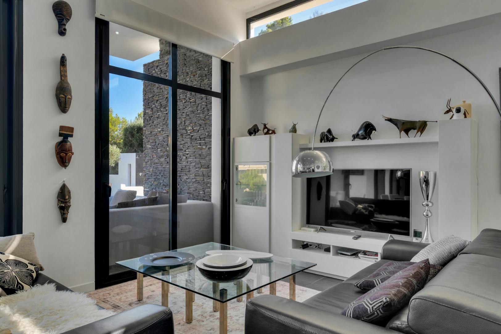 Salon moderne aux hauts plafonds avec menuiseries noires et masques décoratifs. Comprend un lampadaire arc chromé, une table en verre et une vue sur le mur en pierre naturelle.