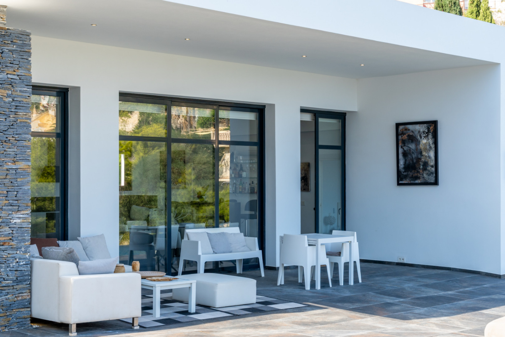 Terrasse couverte avec sol en grès cérame foncé, mur en pierre naturelle et mobilier extérieur blanc minimaliste devant de grandes baies vitrées.