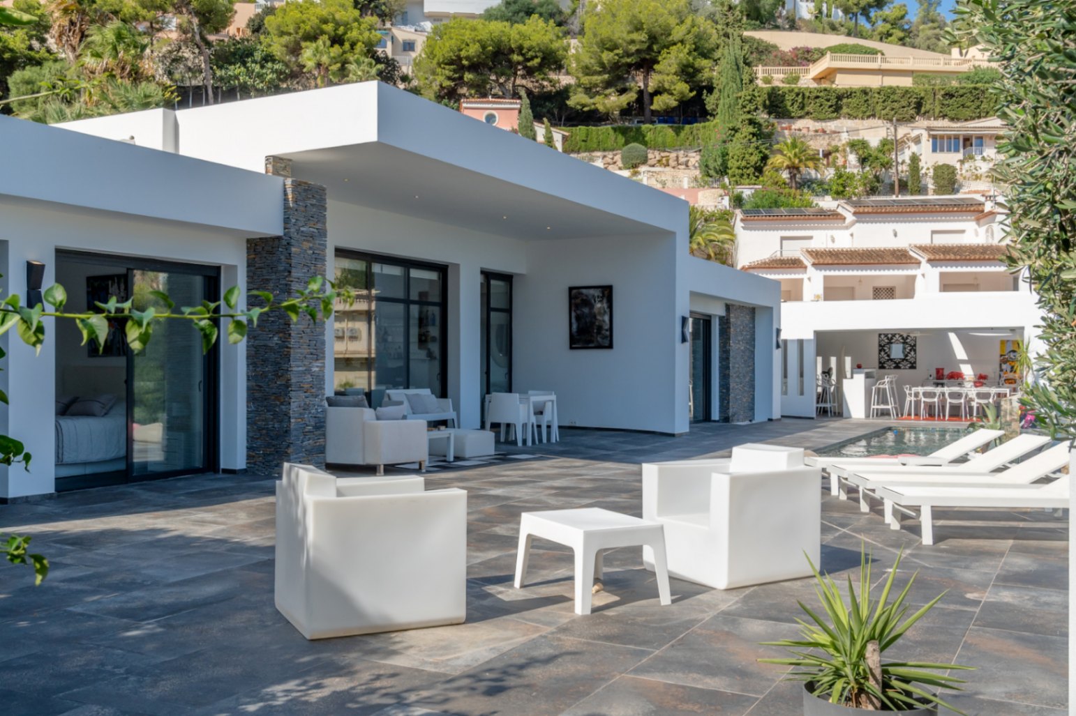 Terrasse extérieure avec sol en pierre foncée, mobilier blanc minimaliste et porche couvert avec piliers en pierre naturelle à côté de la piscine.