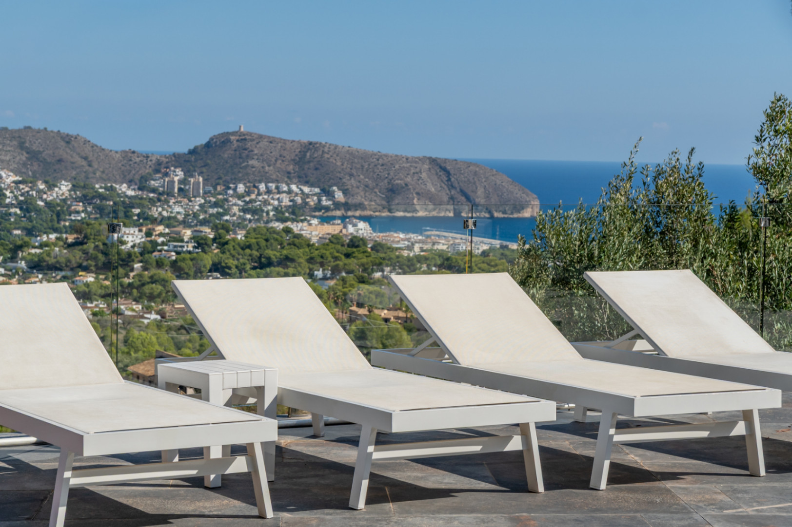 Terrasse solarium équipée de bains de soleil blancs et garde-corps en verre, offrant une vue dégagée sur la mer Méditerranée et Moraira.