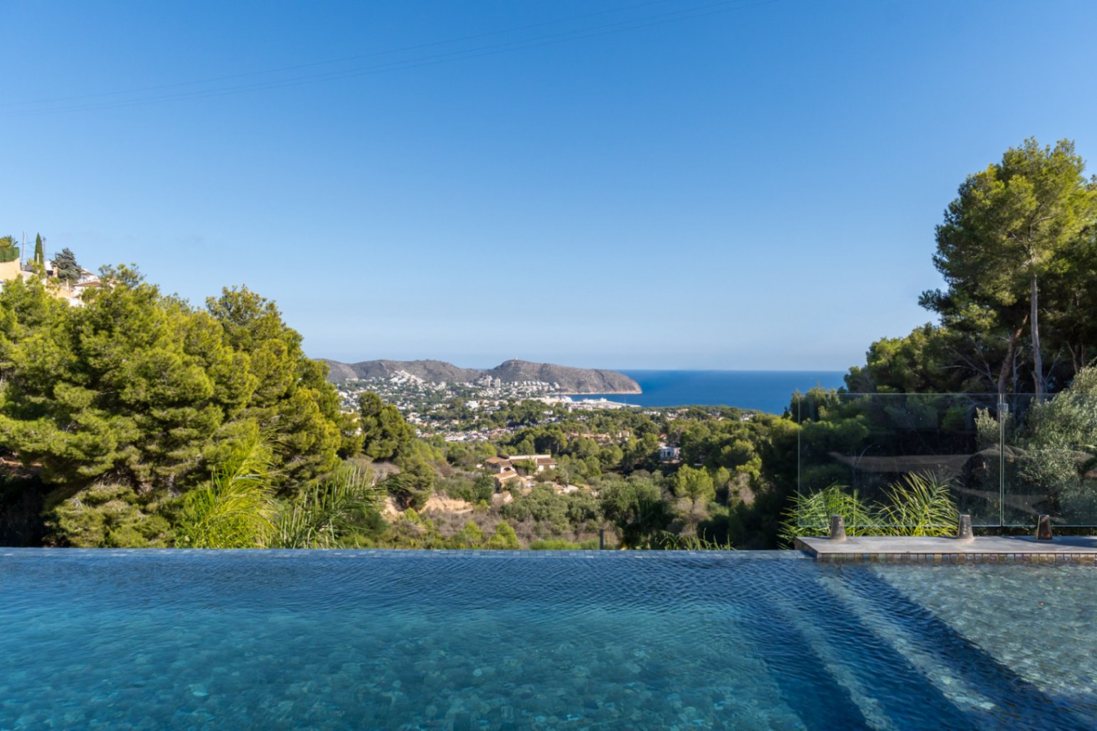 Vues panoramiques sur la mer Méditerranée et la côte de Moraira depuis la piscine à débordement, entourée de pins et de végétation locale.