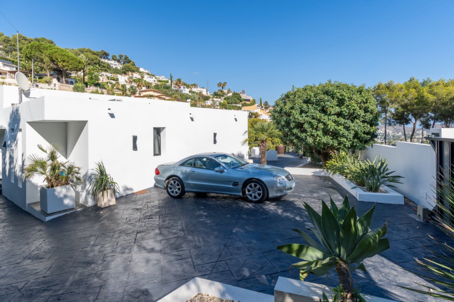Zone d'entrée et parking avec revêtement en béton imprimé foncé, façade minimaliste blanche et jardinières avec végétation méditerranéenne.