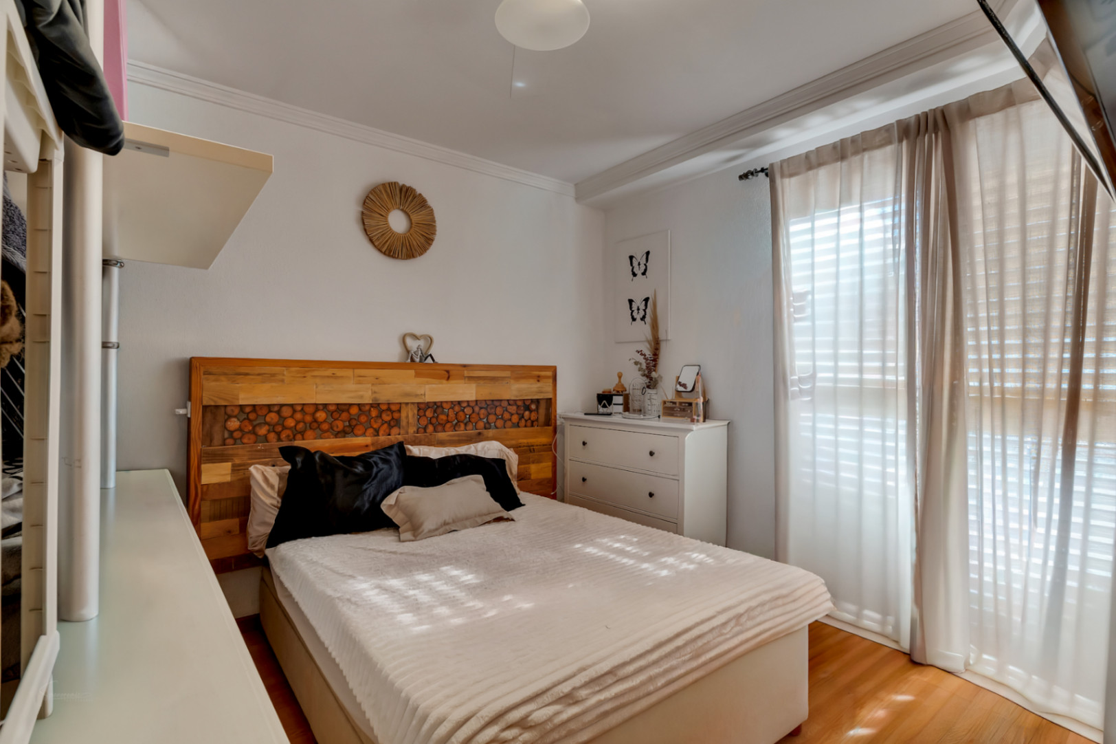 Chambre chaleureuse avec lit double, tête de lit artisanale en bois et sol stratifié. Elle comprend une commode blanche et une grande fenêtre aux rideaux translucides.