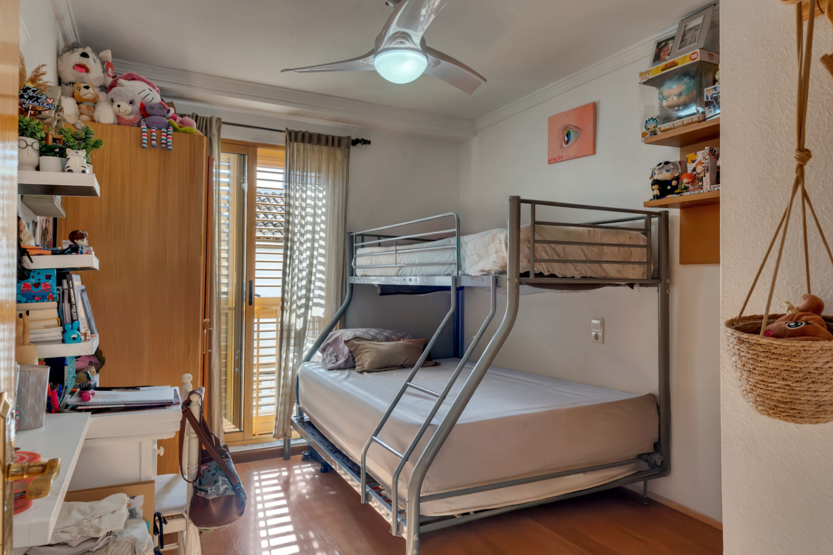 Chambre secondaire avec lits superposés en métal, parquet et ventilateur de plafond. Accès direct à l'extérieur par des baies vitrées équipées de volets traditionnels en bois.